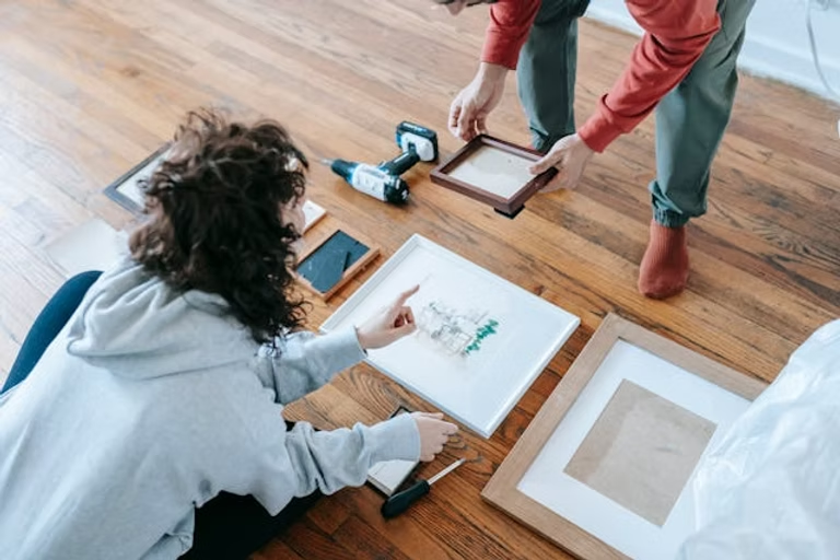 A woman sitting on a wooden floor reviewing artwork while another person holds a picture frame above the tools
