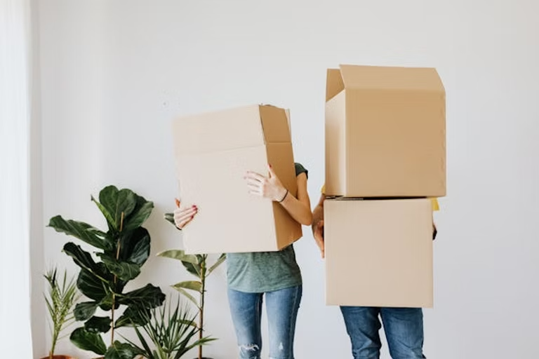 Two people in jeans standing next to each other holding stacked boxes.