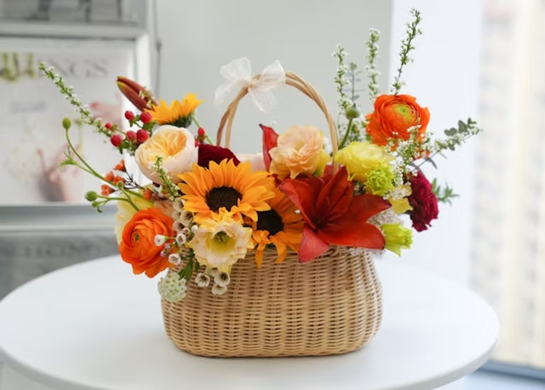 Wicker basket with sunflowers, orange lilies, and roses on a white table. Soft natural light, blurred magazine in the background.