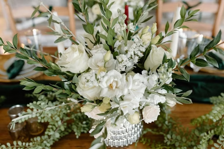 Elegant floral centerpiece with white roses and greenery on a wooden table. Candles and chairs are blurred in the background, creating a cozy effect.
