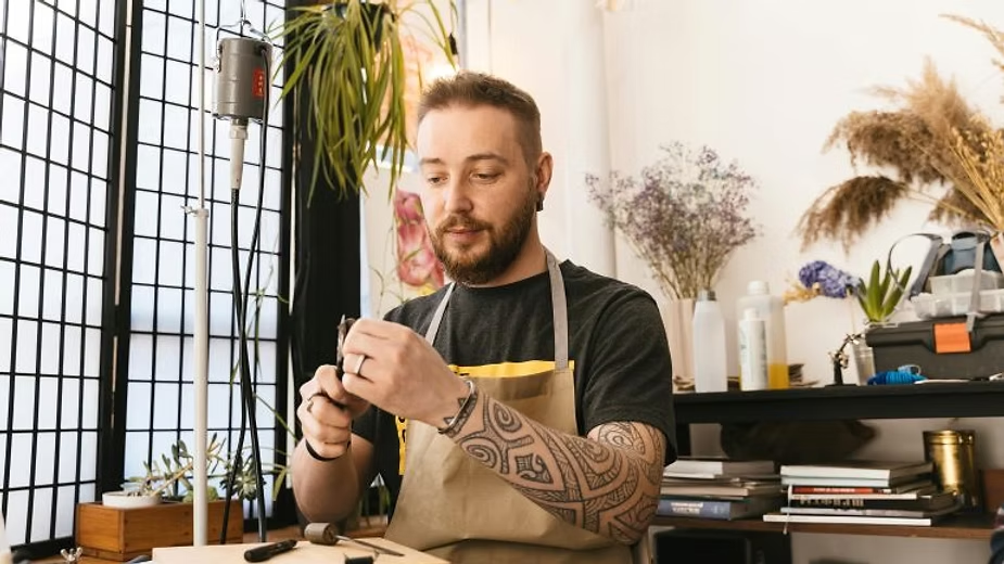 Man with tattoos wearing an apron works on a craft project at a small, organized home workshop.