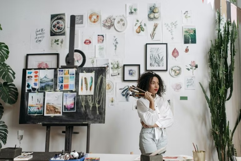 An artist holding brushes stands in a studio surrounded by watercolor art displayed on
the wall.
