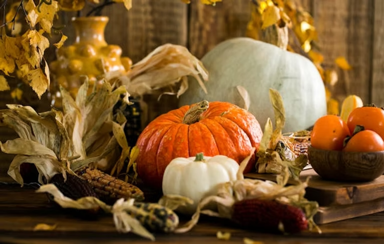 Autumn harvest scene with pumpkins, corn, and persimmons on a wooden table. Yellow leaves, vase, and rustic backdrop create a cozy, warm mood.