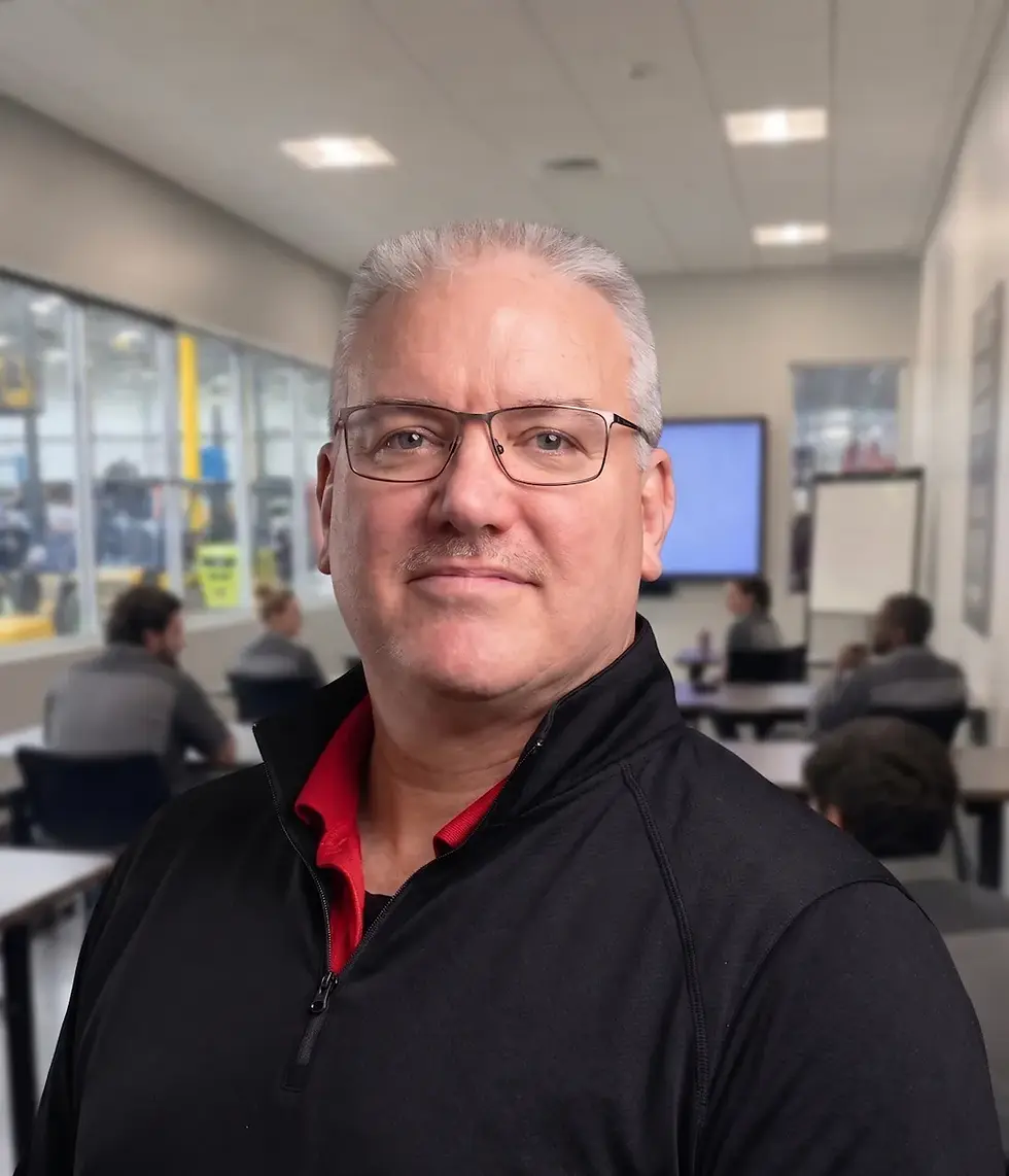 A bust photo of Michael (Mike) J. Brace with a blurred classroom environment with a number of students in their desks, turned towards each other having pleasant conversation. Along one wall is a number of bay windows where a forklift maintained shop is barely visible