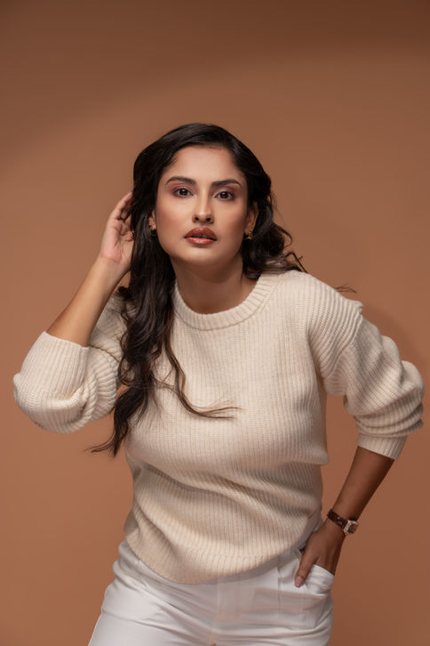 Studio fashion photography of a model wearing a ribbed cream sweater, posing with one hand brushing her hair against a seamless warm brown background.