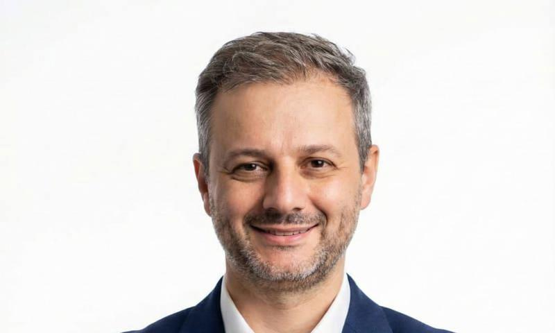 Homem sorridente em traje formal contra fundo branco. Ele exibe expressão amigável e confiante.