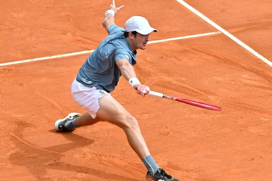 Jogador de tênis se estica para golpear bola em quadra de saibro. Vestido de azul e branco, raquete vermelha em destaque. Ambiente competitivo.
