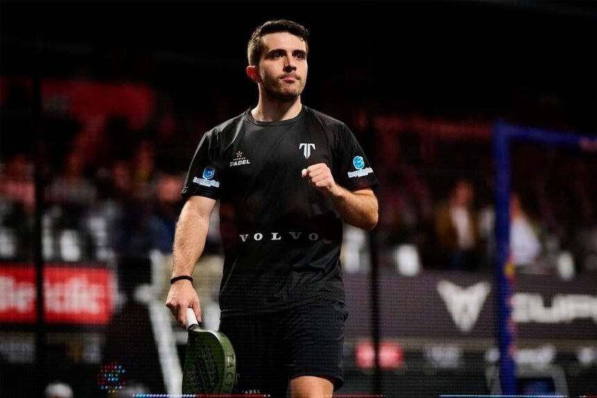 Jogador de padel com uniforme preto, raquete na mão, em quadra coberta. Expressão focada. Logomarcas visíveis na camiseta. Fundo desfocado.