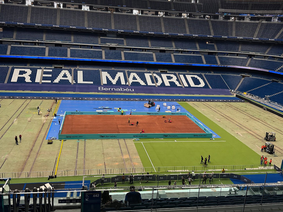 Estádio com quadra de tênis central em montagem. Grama sintética ao redor. Grandes letras "REAL MADRID" no fundo. Pessoas trabalhando.