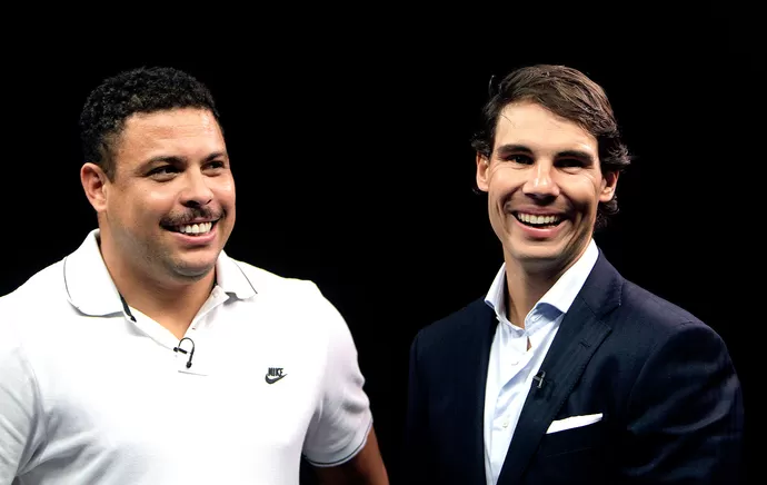 Dois homens sorridentes (Ronaldo Fenômeno e Rafael Nadal), um de camisa branca com logotipo Nike e outro de terno escuro, em fundo preto. Ambos aparentam estar felizes.