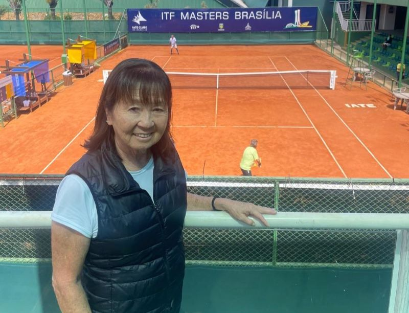 Mulher sorrindo à frente de uma quadra de tênis com piso laranja. Pessoas jogando ao fundo. Placa diz ITF Masters Brasília.