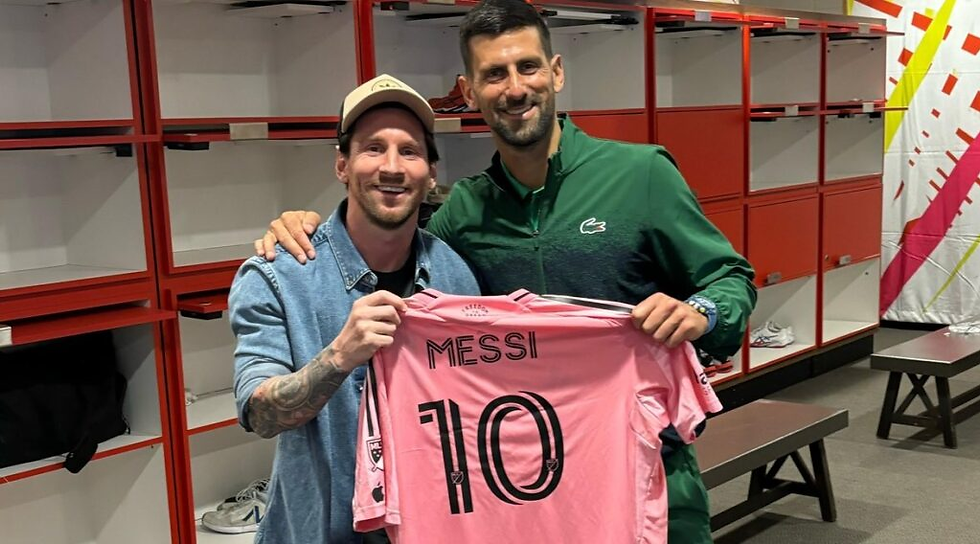 Messi e outro homem sorriem em vestiário, segurando camisa rosa 10 de Messi; armários vermelhos ao fundo.