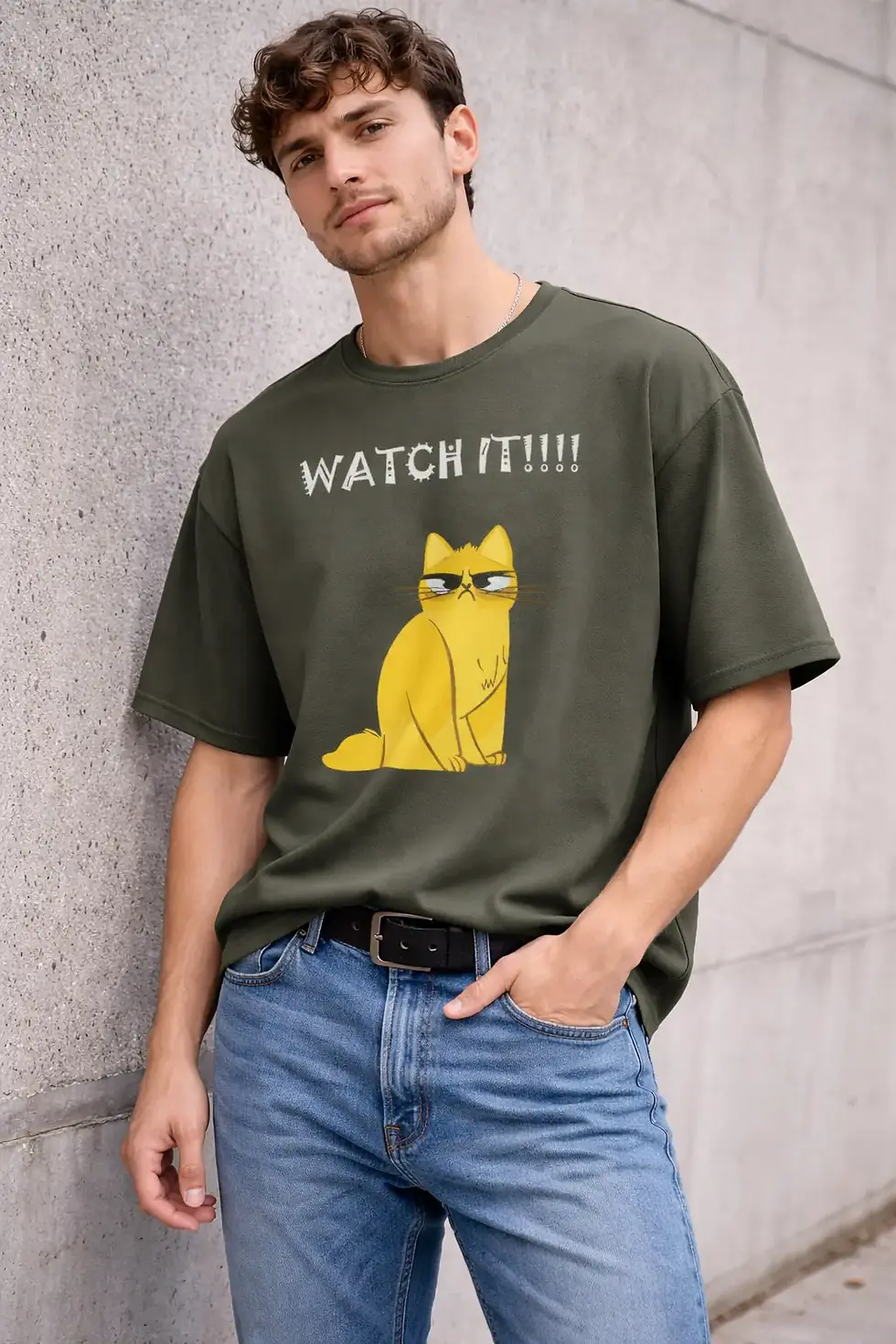 Young man wearing "WATCH IT!!!!" t-shirt with yellow cat, leaning against wall.
