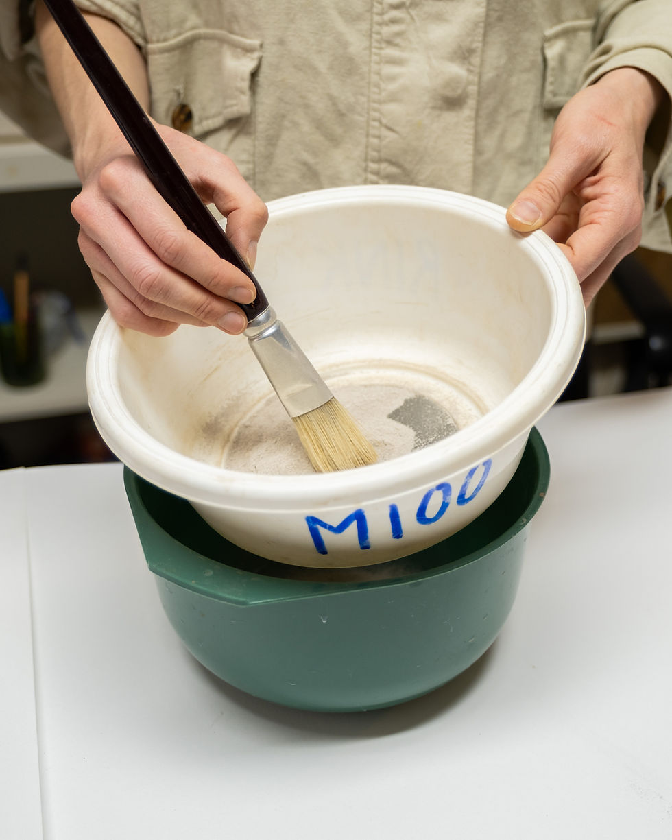 Hand roert glazuurmengsel met penseel in een zeef met label M100 tijdens glazuurbereiding bij Urban Glaze Studio