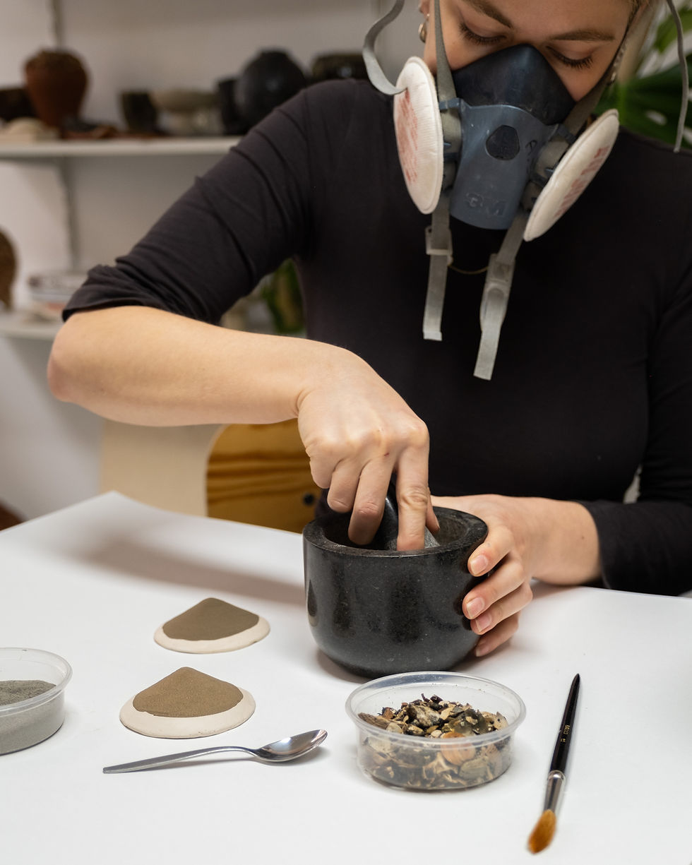 Rink Schelling van Urban Glaze Studio maalt glazuuringrediënten in stenen vijzel met stofmasker op, omringd door keramische tegelproeven