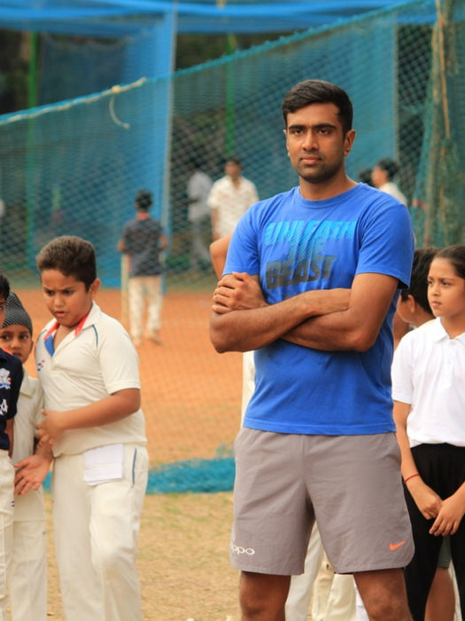 Ravichandran Ashwin standing with arms crossed, observing a group of young cricketers during a practice session.