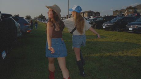 Two friends in casual festival outfits walking through the grassy parking lot of a car boot sale at sunset, smiling and carefree.