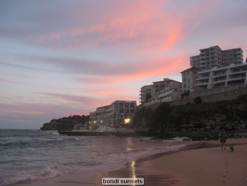 Beach at sunset with buildings and pink sky.