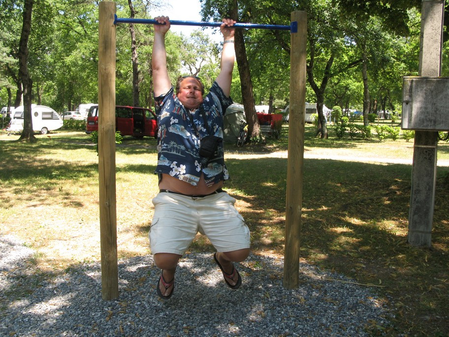 Man hanging from pull-up bar outdoors, wearing casual clothes.