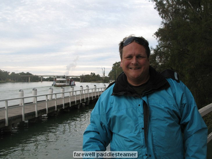 Man in blue jacket by river with paddle steamer.
