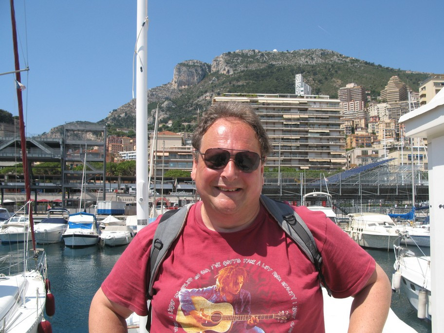 Man smiling at marina with boats and mountains.