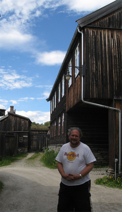 Man standing by rustic wooden buildings under blue sky.
