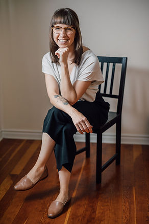Angie sitting on a black chair wearing black pants, white shirt and glasses with shoulder-length brown hair with bangs