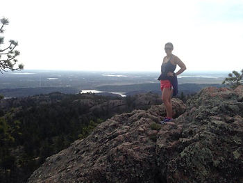 This picture starts with a rock in the front of the picture with Katie (a female) standing on top of the rock. Behind the rock is an overlook of grass areas and small lakes. To the left of the picture is a small tree that is slightly in the picture.