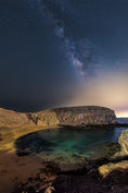 A breathtaking night view of Playa Papagayo, Lanzarote, captured under the brilliance of the Milky Way. The golden curves of the secluded beach and rugged volcanic cliffs meet the calm Atlantic waters, while the star-studded sky arches overhead in dazzling detail.