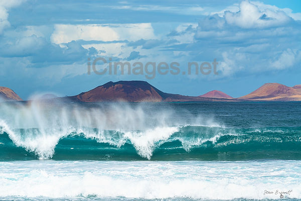 This photograph captures the rolling waves of the Atlantic Ocean as seen from Playa Famara, with the stunning silhouette of La Graciosa island in the background. The scene showcases the untamed beauty of Lanzarote’s northwest coast, where powerful waves crash onto the golden sandy shore, leaving behind delicate white foam patterns. In the distance, La Graciosa’s rugged landscape emerges from the horizon, bathed in soft light. The contrast between the dynamic ocean and the serene island creates a breathtaking and timeless coastal view.