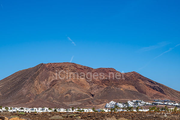 A stunning view of Montaña Roja, a striking reddish-brown volcanic cone in Playa Blanca, Lanzarote, set against a brilliant blue sky. The rugged slopes of the volcano contrast with the arid landscape. The scene is bathed in warm sunlight, highlighting the unique textures of the volcanic terrain.