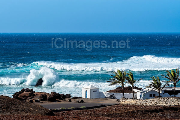 A dramatic photograph of El Golfo, a charming fishing village in Lanzarote, on a windy day. Towering waves crash against the rugged beach, sending white spray into the air, while the wild Atlantic Ocean showcases its raw power against the volcanic coastline.