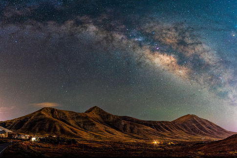 This stunning photograph showcases the Milky Way arching in a brilliant display over the rugged Femes Mountains of Lanzarote. The night sky becomes a celestial canvas, illuminated by countless stars and the glowing galactic core radiating with hues of blue, purple, and pink. The sweeping Milky Way arch creates a breathtaking contrast against the dark volcanic silhouette of the peaks, capturing both the serenity and otherworldly beauty of Lanzarote under a starlit sky.