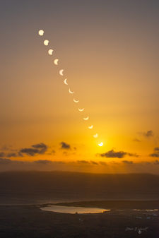 A dramatic sequence of solar eclipse (2026) phases unfolds across a glowing orange sky, with the sun gradually disappearing and re-emerging through the celestial progression. The warm amber and golden tones create a striking, atmospheric backdrop, while the silhouettes of the landscape add depth and contrast. At the bottom of the photograph, the distinctive salt pans of Salinas de Janubio stretch across the foreground, their geometric pools reflecting the rich colors of the sky.