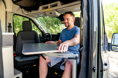 Person sitting at a Lagun table inside a Ford Transit camper van with swivel seating, kitchen galley, and front cab workspace.