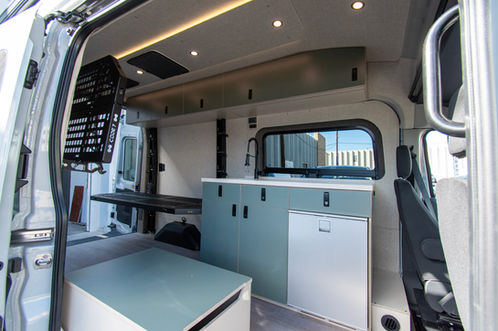 Voyager Ford Transit camper van kitchen galley with bench storage, refrigerator, overhead cabinets, side window, and Code 7 adjustable bed system.