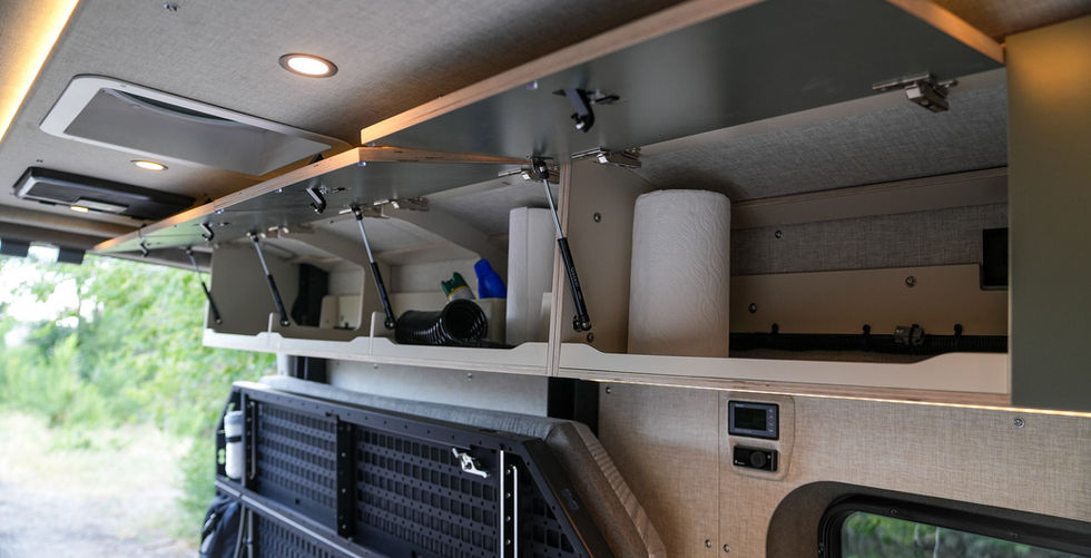 Open upper cabinets inside a Voyager Ford Transit camper van with kitchen storage, gas strut supports, Code 7 bed platform, and galley below.