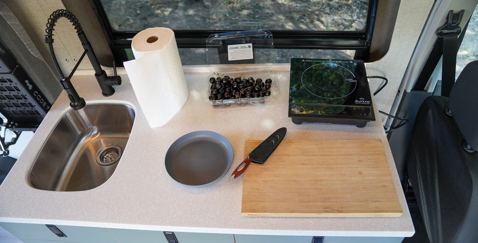Voyager Ford Transit camper van kitchen counter with portable induction cooktop, sink, cutting board, refrigerator, vented window, and meal prep setup.