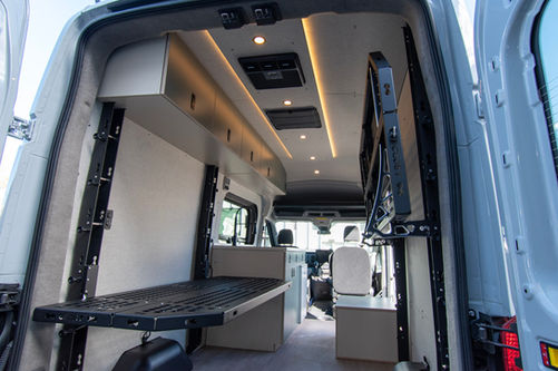Rear interior of a Voyager Ford Transit camper van with Code 7 adjustable bed platform, overhead cabinets, LED ceiling lights, wall panels, and gear space.
