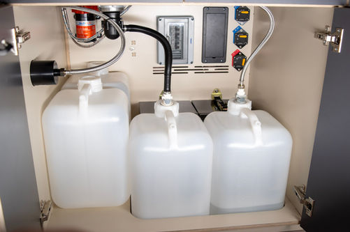 Water and electrical system under a Voyager camper van kitchen galley with removable water jugs, plumbing, pump, breaker panel, and main power switches.