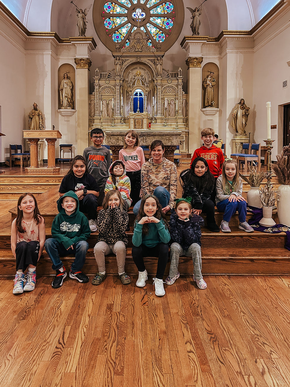 A group of 14 people, including children, sit on wooden steps in a church with ornate statues and stained glass. They are smiling and relaxed.