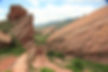 Red rock formations rise against a cloudy blue sky at Red Rocks Park, surrounded by greenery and distant mountains.