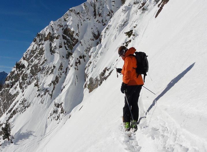 Climber in blue jacket and helmet scales rocky cliff with snowy mountains and a lake below, conveying focus and determination.