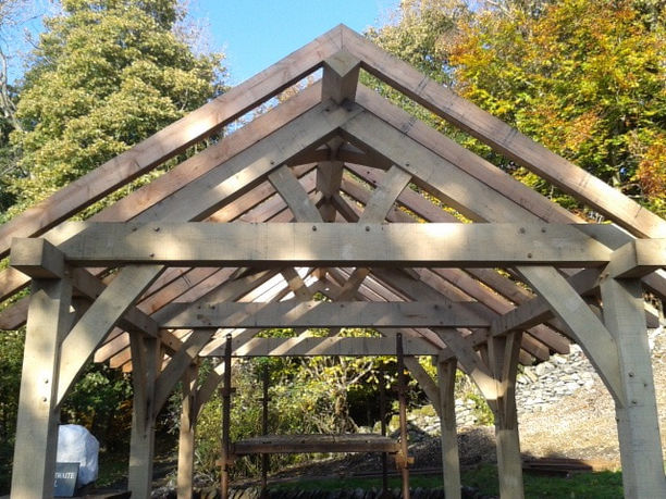 Timber frame car port under construction with oak beams forming a pitched roof in a woodland setting.