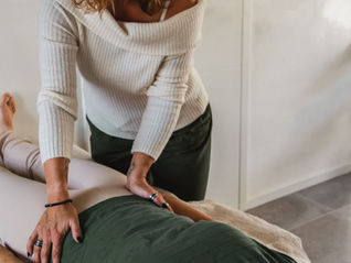 Twee vrouwen in lichte kamer, één geeft massage aan de ander op een tafel. De masseuse draagt een witte trui, de ander groen shirt. Ontspannen sfeer. Energiewerk nodigt dat proces uit, in rust en afstemming.