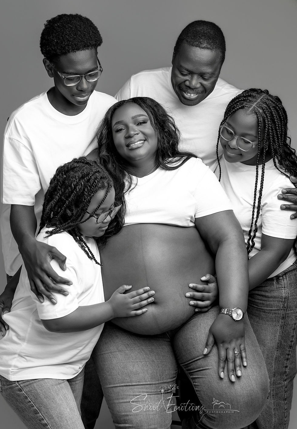 Famille souriante en noir et blanc, entourant une femme enceinte, portant des t-shirts blancs. Les enfants touchent tendrement son ventre.