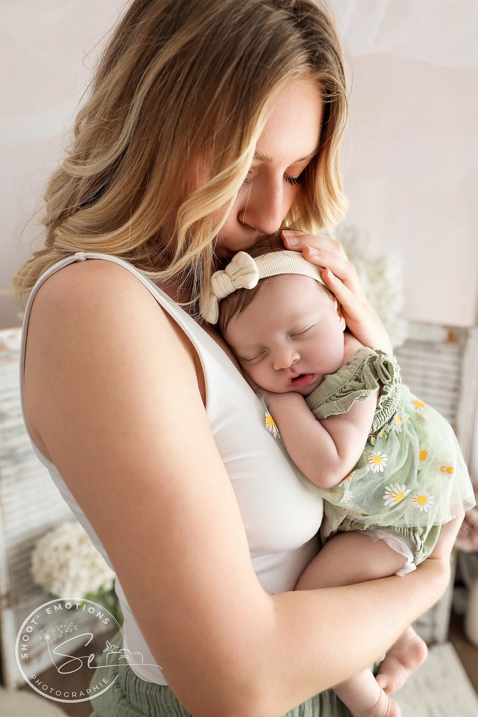 Mère embrassant un bébé endormi, dans un décor lumineux; robe verte à fleurs, logo Shoott Emotions Photographie en bas.