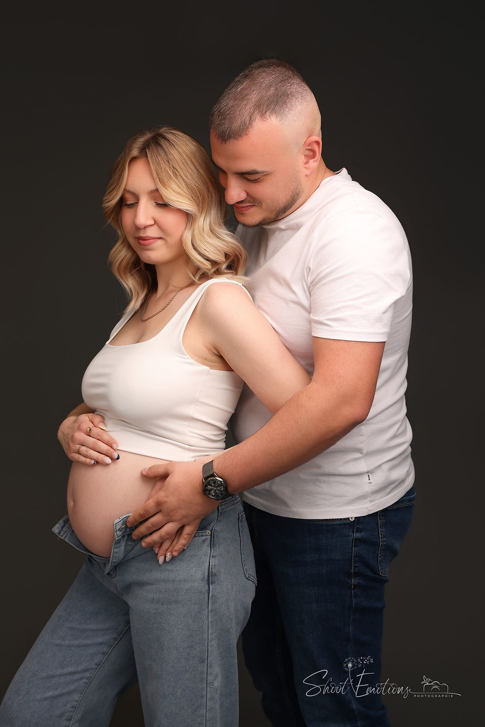 Couple souriant, homme tenant le ventre de la femme enceinte. Fond sombre, vêtements blancs et jeans. Atmosphère tendre et joyeuse.