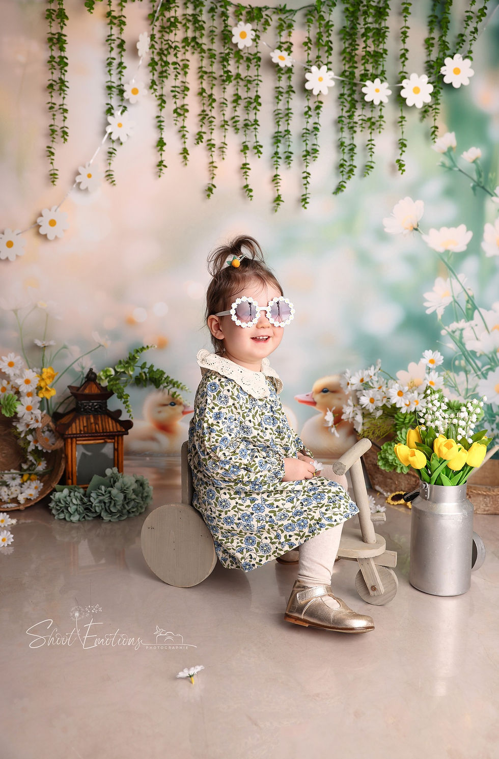 Petite fille souriante avec lunettes de soleil fleuries, robe à motifs bleus, assise sur un tricycle en bois, décor floral et fond verdoyant.