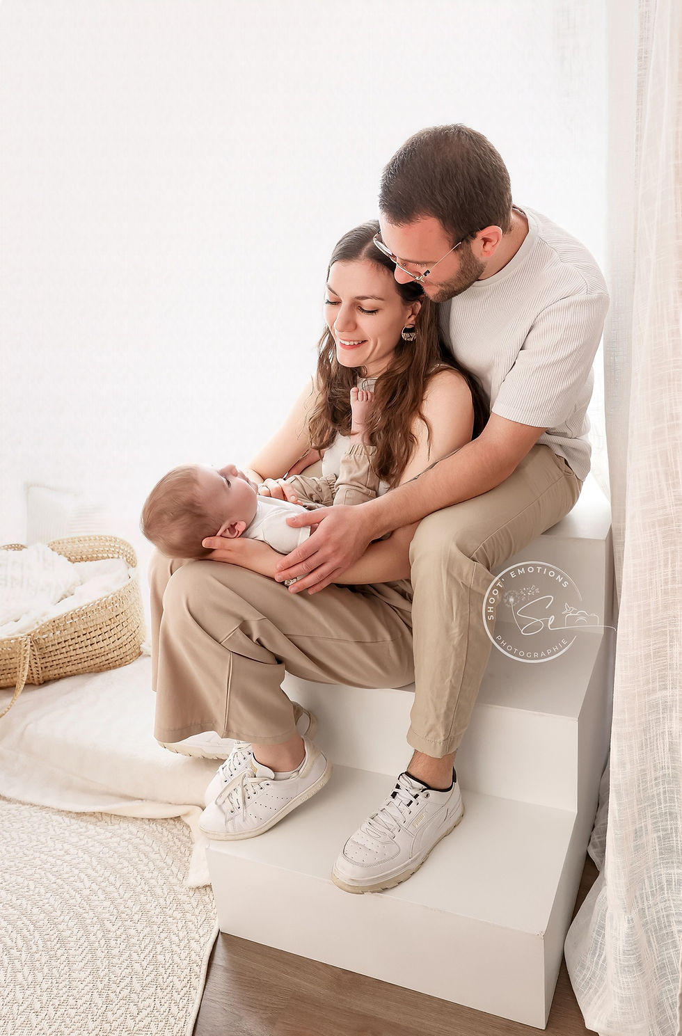 Un couple souriant tient un bébé dans un cadre lumineux avec des tons neutres. Ambiance douce et chaleureuse.